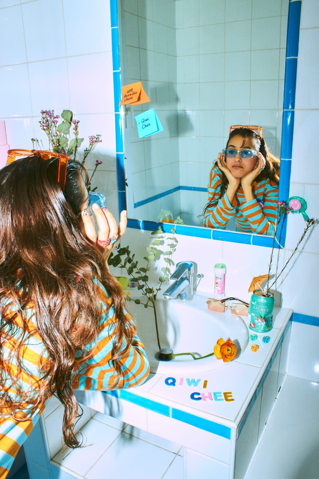 Qiwi Chee de dos devant un miroir, son reflet le menton posé dans les mains ; sur la glace, deux post-it portent les mots « une dernière chose » et « Qiwi Chee », et les lettres autocollantes QIWI CHEE sont collées sur le lavabo.